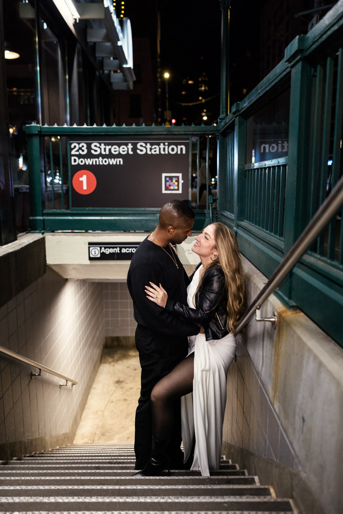 A couple embraces and laughs on stairs at the 23 Street subway station entrance at night.