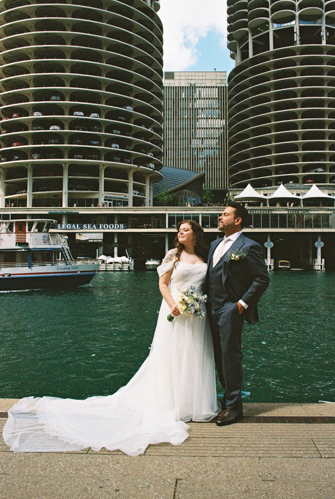 Bride and groom pose by the river, modern city buildings and a boat in the background.