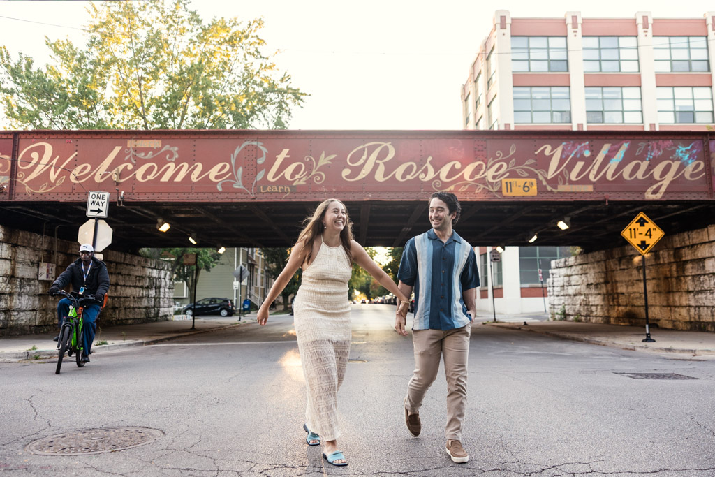 Engaged couple laughs while holding hands and crossing the street in front of an overpass with 