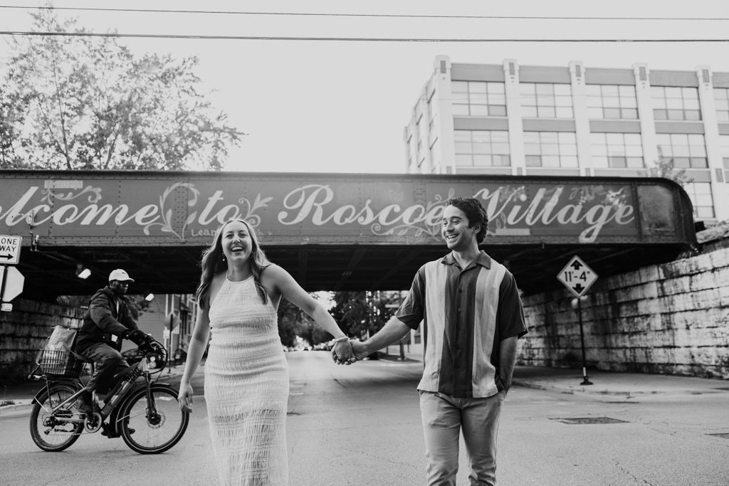 Black and white photo of engaged couple laughing while holding hands and crossing the street in front of an overpass with 