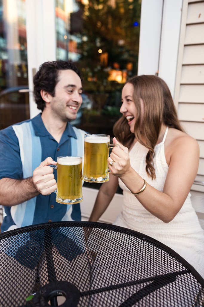 Happy couple clinking beer mugs while sitting at an outdoor table at Four Moons Tavern during their Roscoe Village engagement session