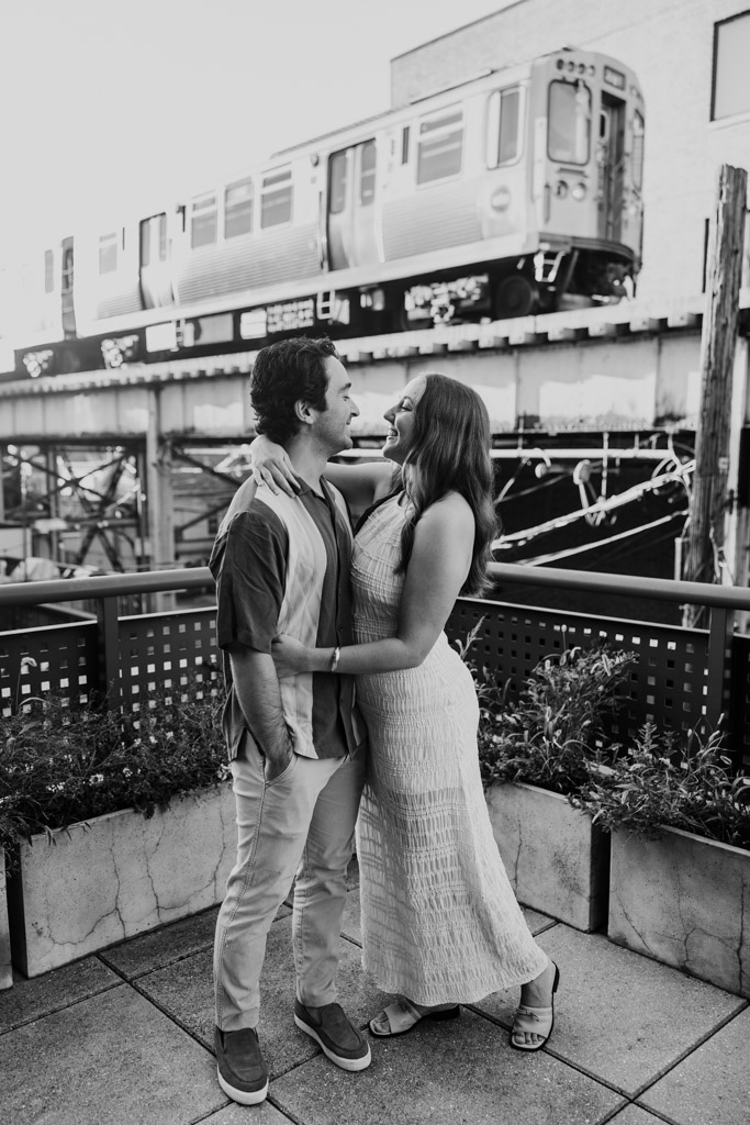 Black and white photo of couple embracing and smiling at each other on a rooftop during their Roscoe Village engagement session with a train passing by in the background
