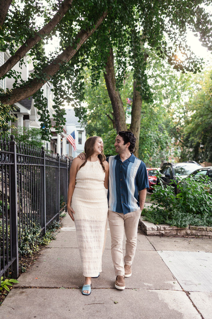 Happy couple walking arm in arm on a tree-lined Roscoe Village sidewalk during their summer engagement session