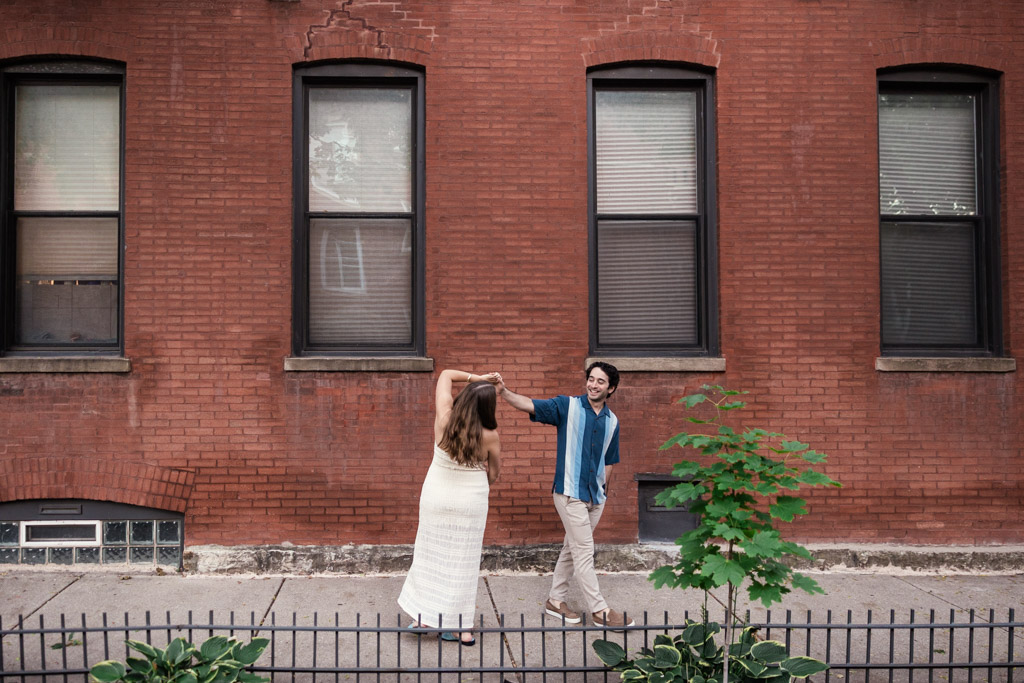 Engaged couple dances on the sidewalk in front of a red brick building with three large windows during their Roscoe Village engagement session