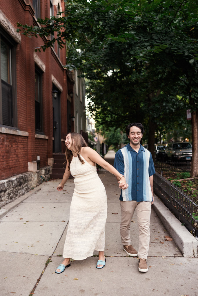 Happy engaged couple holds hands while walking on a city sidewalk lined with trees and brick buildings during Roscoe Village engagement session