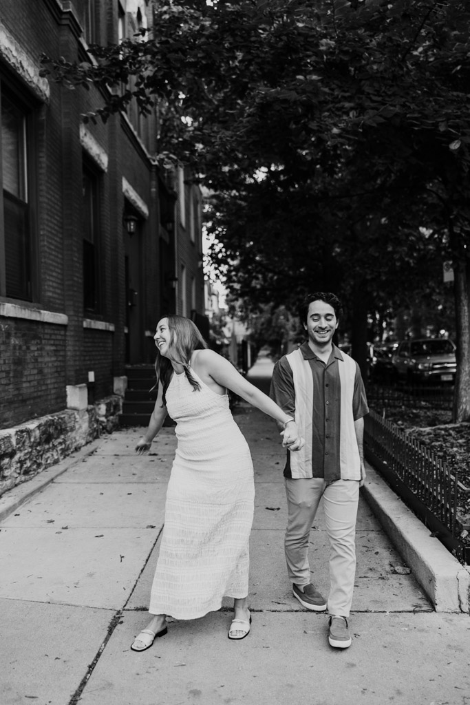 Black and white photo of happy couple holding hands and walking on a sidewalk in Roscoe Village, surrounded by trees and buildings, during their summer engagement session