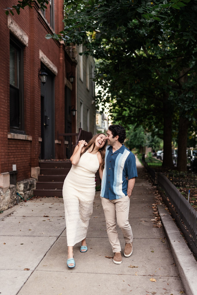 Happy engaged couple walks down a tree-lined sidewalk in Roscoe Village, laughing and enjoying each other's company during their summer engagement session