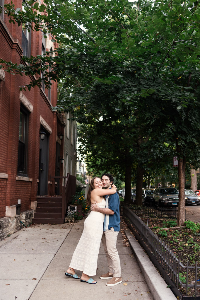 Couple hugs and smiles on a tree-lined city sidewalk beside a brick building, during their Roscoe Village engagement session