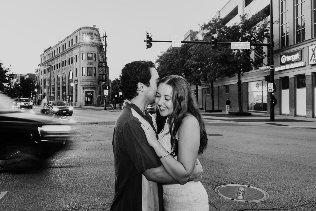 Black and white photo of couple standing on a Roscoe Village street corner as cars pass by in the background during their summer engagement session