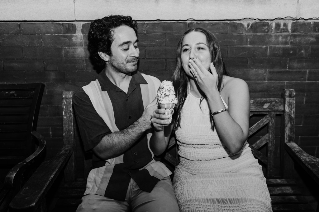 A man holds an ice cream cone as a woman beside him laughs, covering her mouth, seated on a bench during their Roscoe Village engagement.