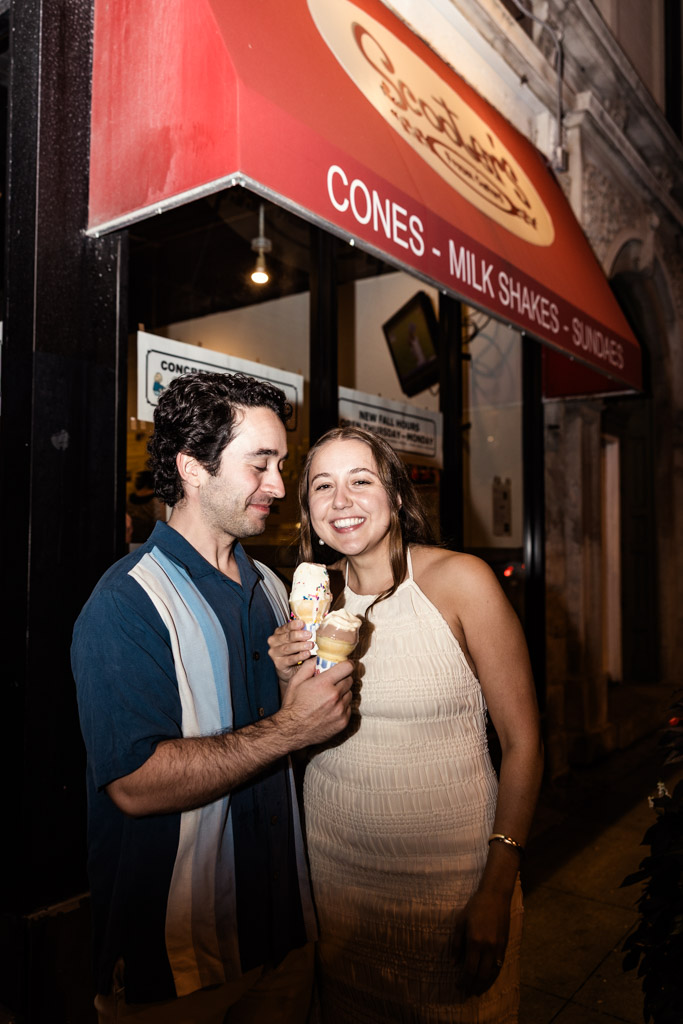 Happy couple stands outside Scooters Frozen Custard in Roscoe Village, holding ice cream cones together at night during their summer engagement session