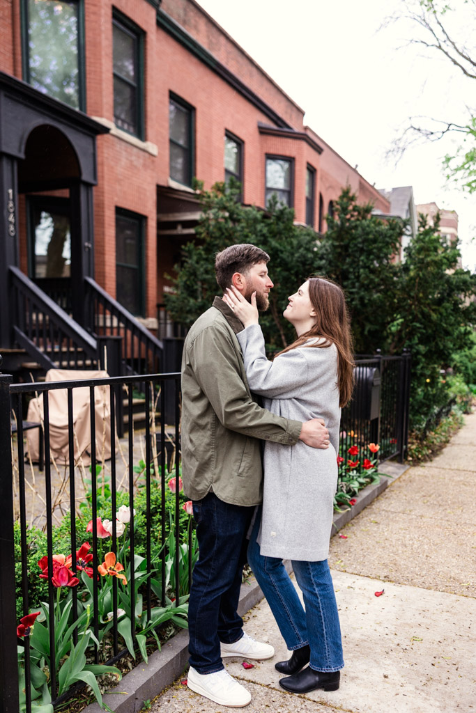 Engaged couple holds each other on a sidewalk in front of brick townhouses and blooming spring flowers