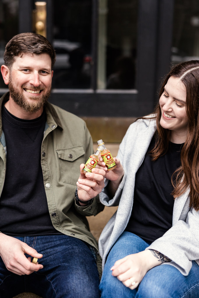 A man and woman smiling and clinking small bottles together while sitting outside, celebrating their sunrise engagement.