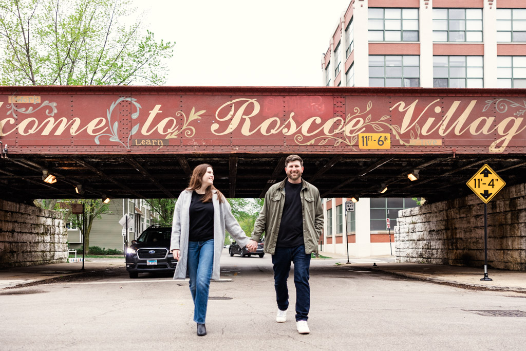 A couple holds hands while walking under a “Welcome to Roscoe Village” rail bridge during their sunrise engagement session