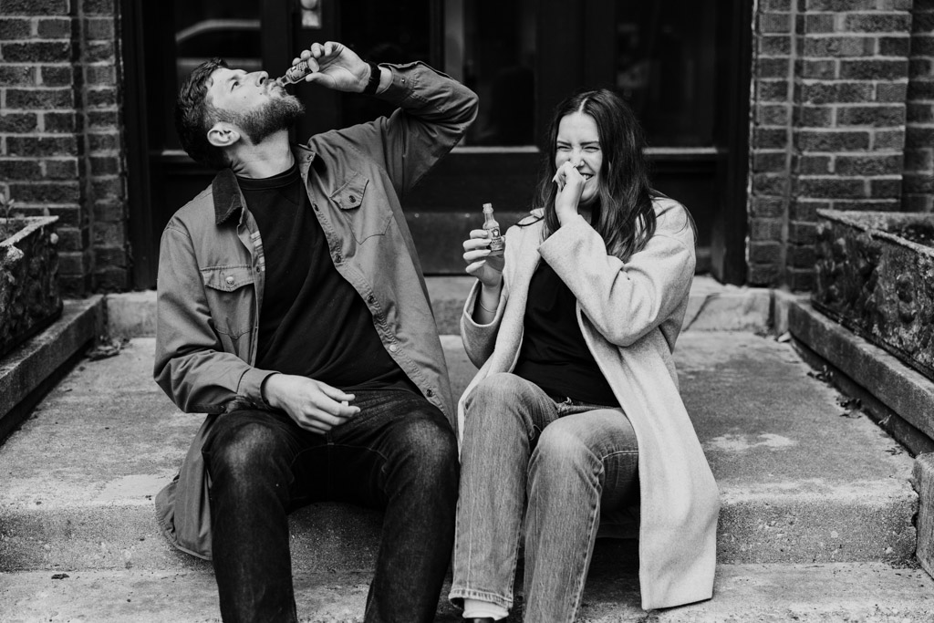 Engaged couple sits on the steps to their apartment building in Roscoe Village, laughing and drinking small bottles of Malört during their sunrise engagement session