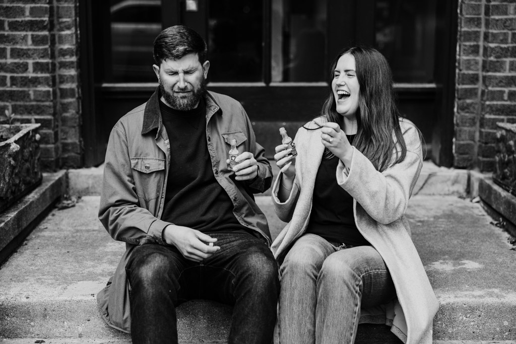 Black and white photo of man and woman sitting on the steps to their apartment building at sunrise drinking Malört; she laughs while he reacts with a grimace