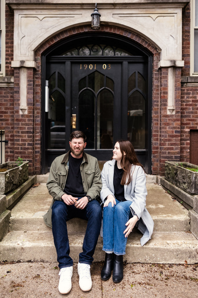 A man and woman sit on steps outside a brick building with black double doors during their sunrise engagement session