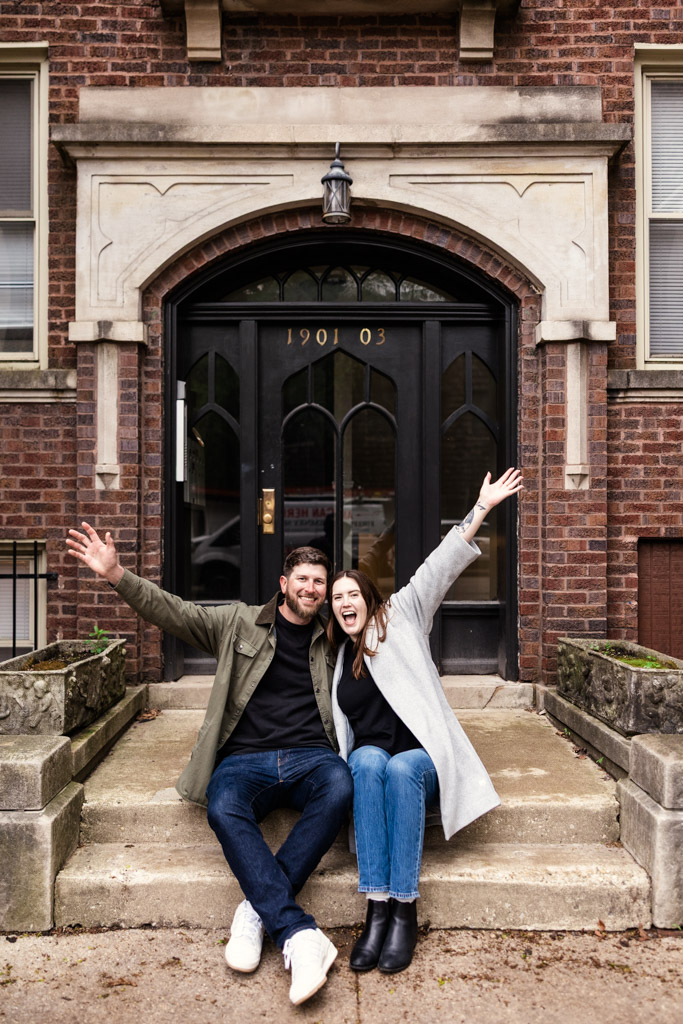 Happy engaged couple sits on steps in front of a brick building, smiling with arms raised in excitement during their Roscoe Village sunrise engagement session