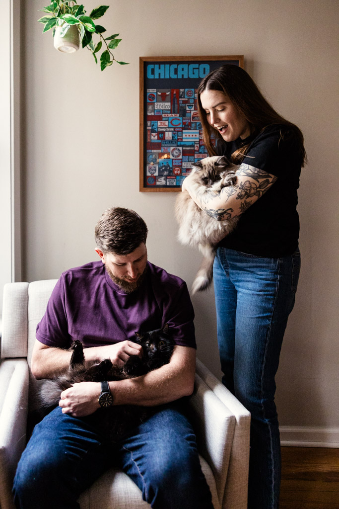 A man sits petting a black cat while a woman stands holding a fluffy cat near him inside their Roscoe Village home during their sunrise engagement session