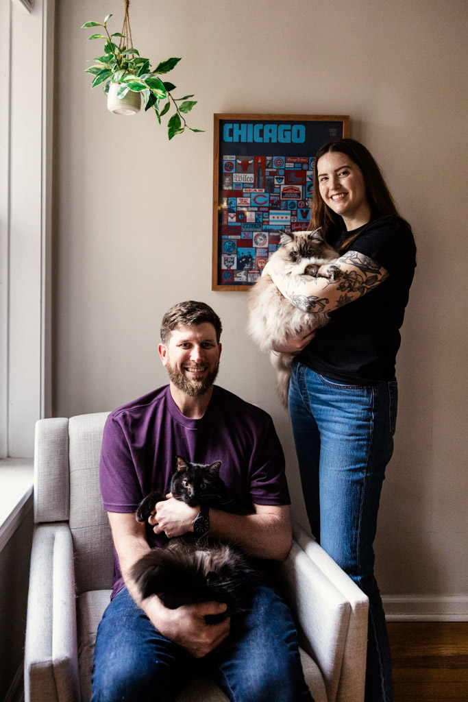 A smiling man sits holding a black cat; a woman stands beside him with a fluffy cat, both glowing like a sunrise engagement, with a Chicago poster behind.
