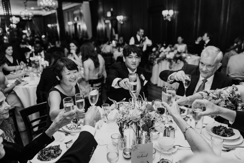 Black and white photo of guests raise glasses in a toast around a decorated table in Salvatore's wedding reception space