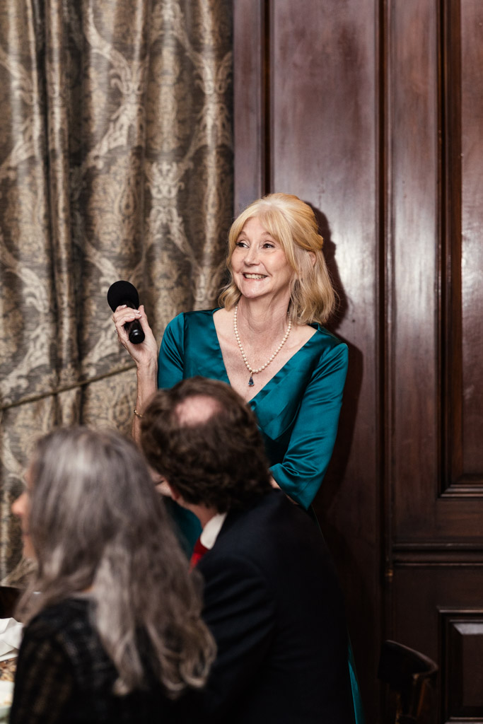 Groom's mother in a teal dress smiles while holding a microphone during wedding reception at Salvatore's