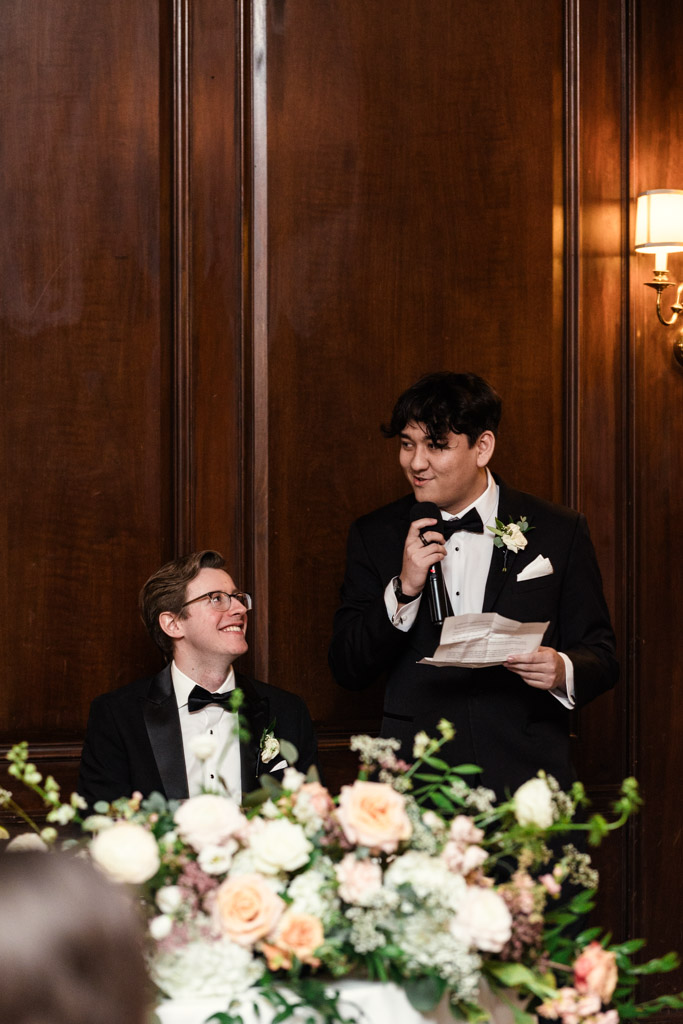 Groom listens to bride's brother giving speech by a flower-filled head table at Salvatore's