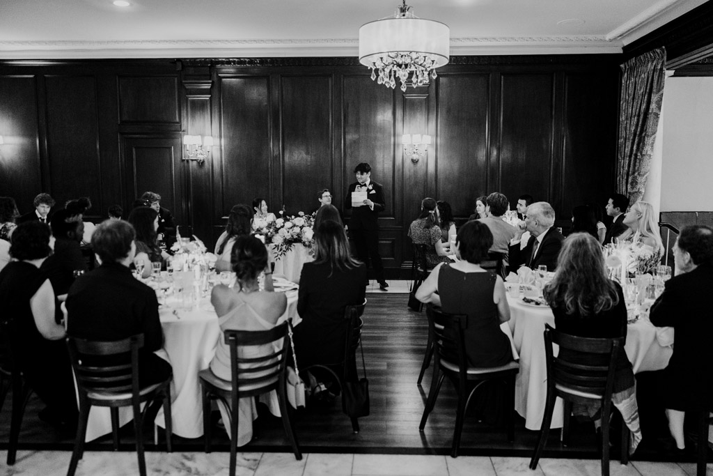 Black and white photo of bride's brother giving a speech during wedding reception at Salvatore's, with guests seated at round tables in a wood-paneled room