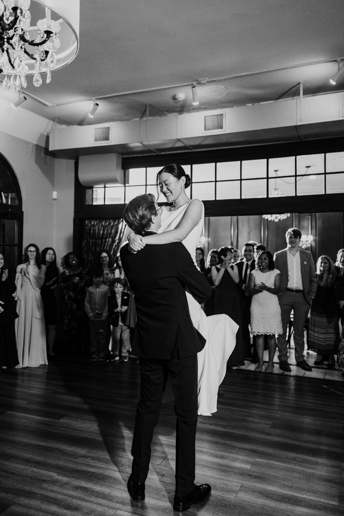 Black and white photo of groom lifting his smiling bride as guests watch during their first dance at Salvatore's
