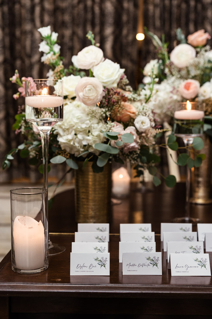 Wedding place cards on a table at Salvatore's, with floral centerpieces and lit candles in glass holders glowing softly in the background