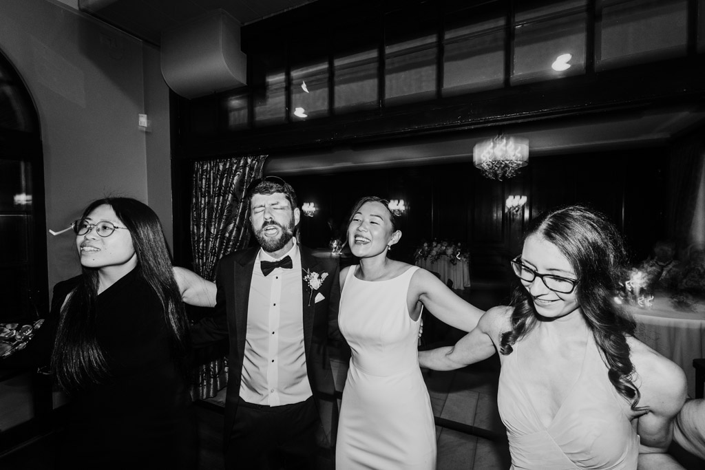 Black and white photo of bride and guests smile and link arms while dancing during wedding reception at Salvatore's in Chicago