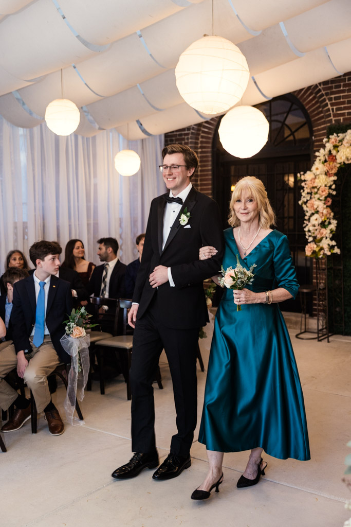 Groom in a tuxedo walks arm-in-arm with ahis mother in a teal dress at Salvatore's wedding ceremony