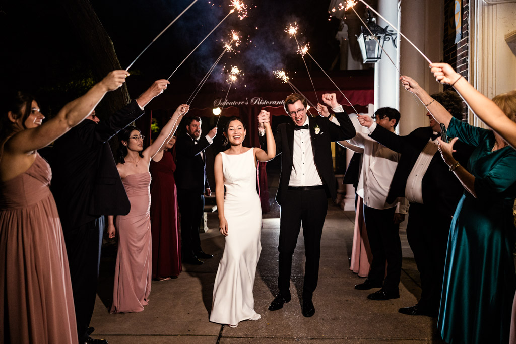 Bride and groom walk under sparklers held by friends, smiling and celebrating at night after their Salvatore's wedding