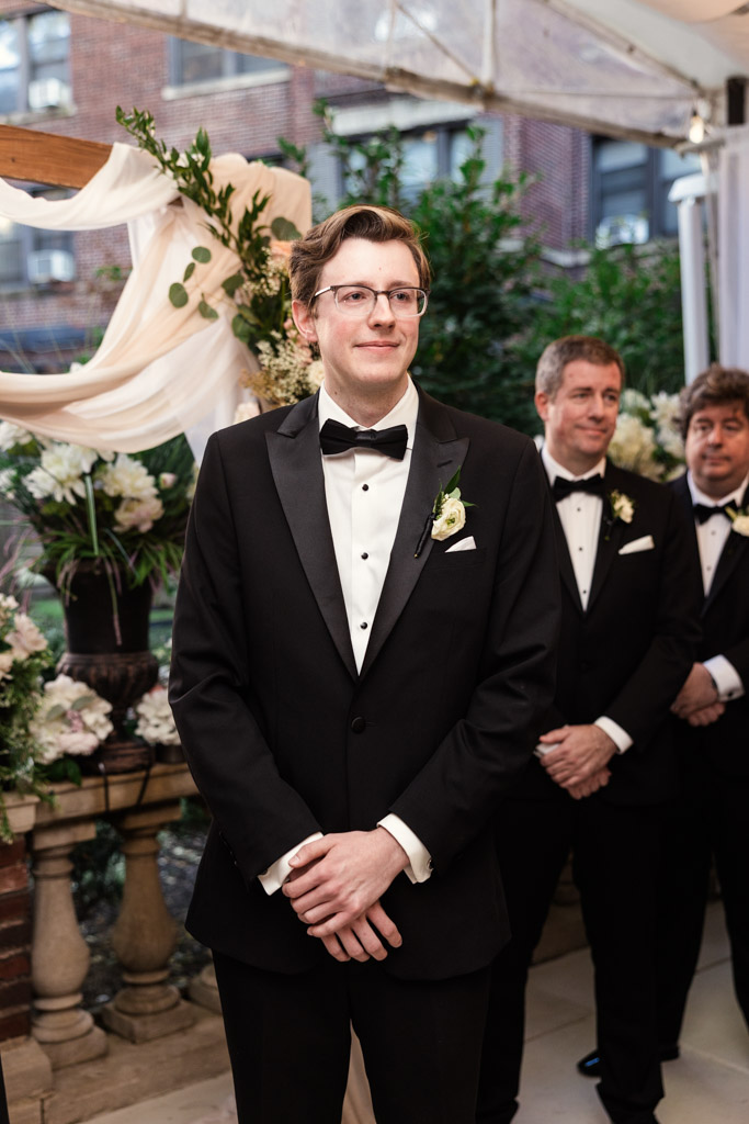 Groom in a black tuxedo stands smiling indoors at Salvatore's, with groomsmen and floral decorations behind him