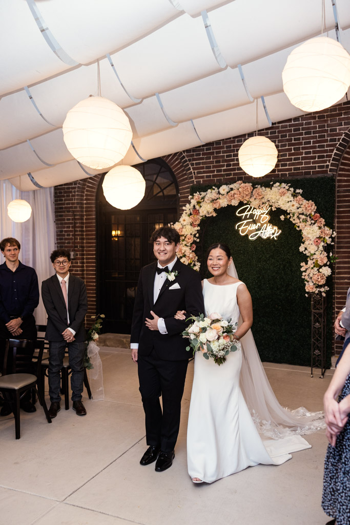 Bride and her brother smiling, walking down the aisle under white lights and a floral wedding arch as they enter wedding ceremony at Salvatore's