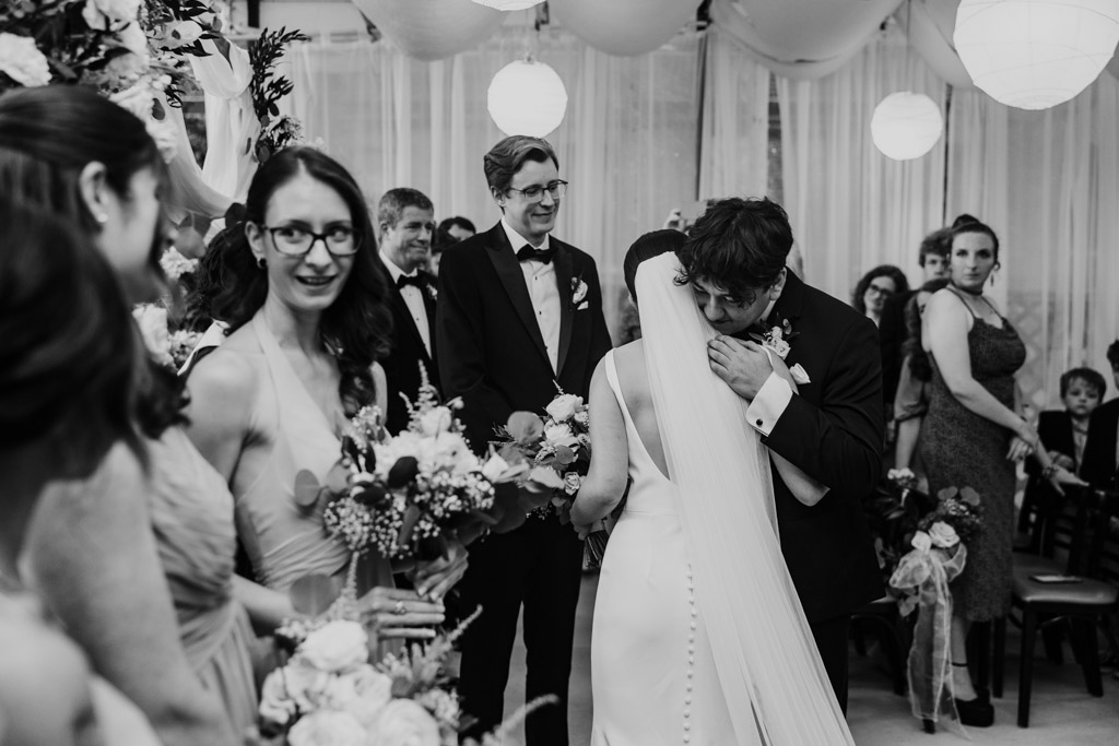 Black and white photo of bride hugging her brother at her wedding ceremony at Salvatore's, surrounded by smiling guests and bridesmaids