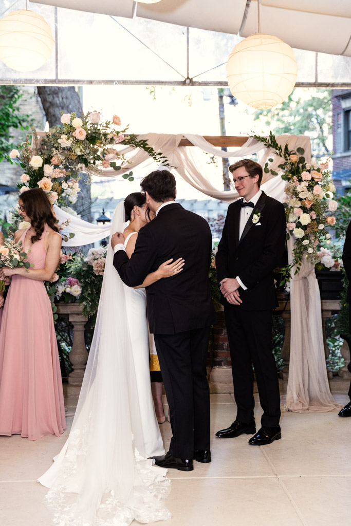 Bride and her brother hug under a floral arch while the groom looks on at their Salvatore's wedding ceremony