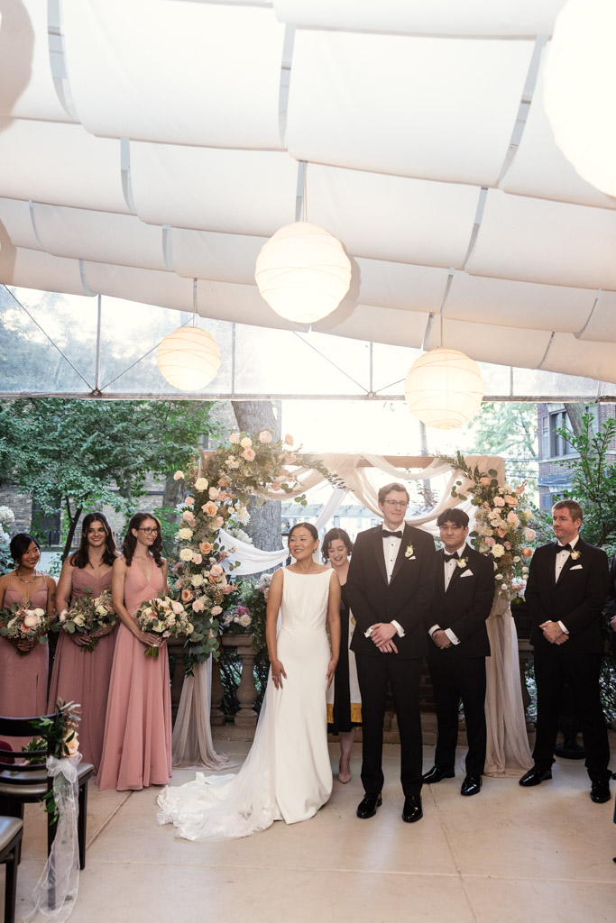 A bride and groom stand at their wedding ceremony at Salvatore's, surrounded by their bridal party under a floral arch
