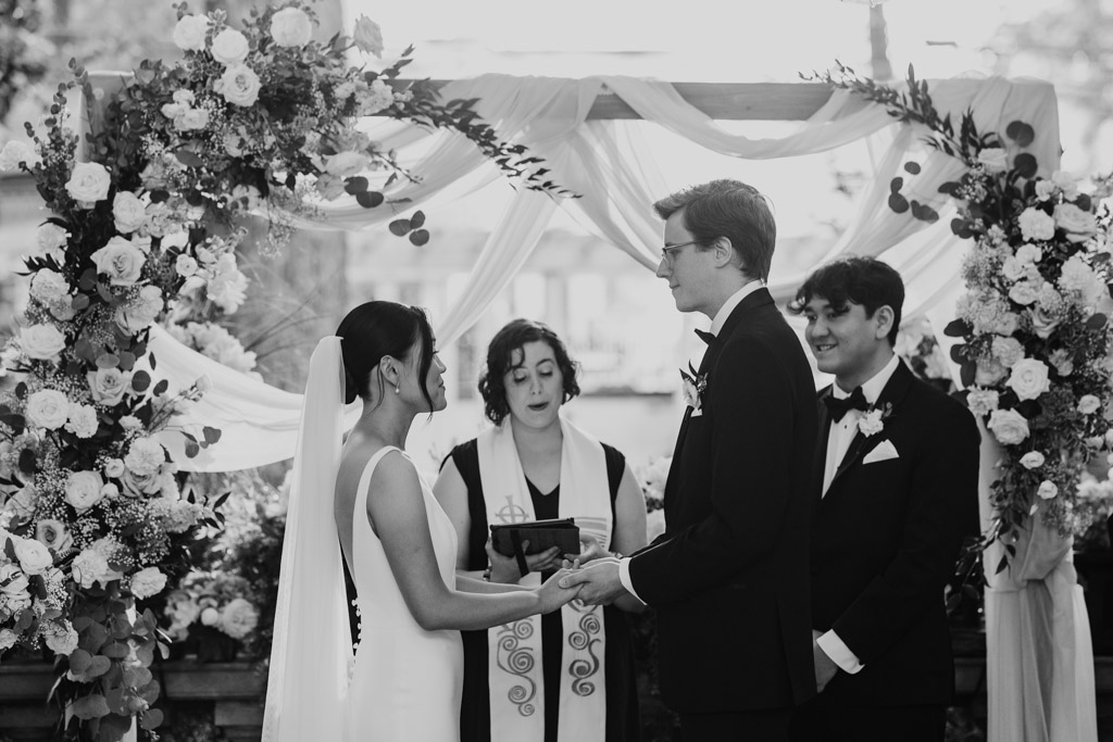 Black and white photo of bride and groom holding hands while officiant speaks at their wedding ceremony at Salvatore’s, standing under a floral arch