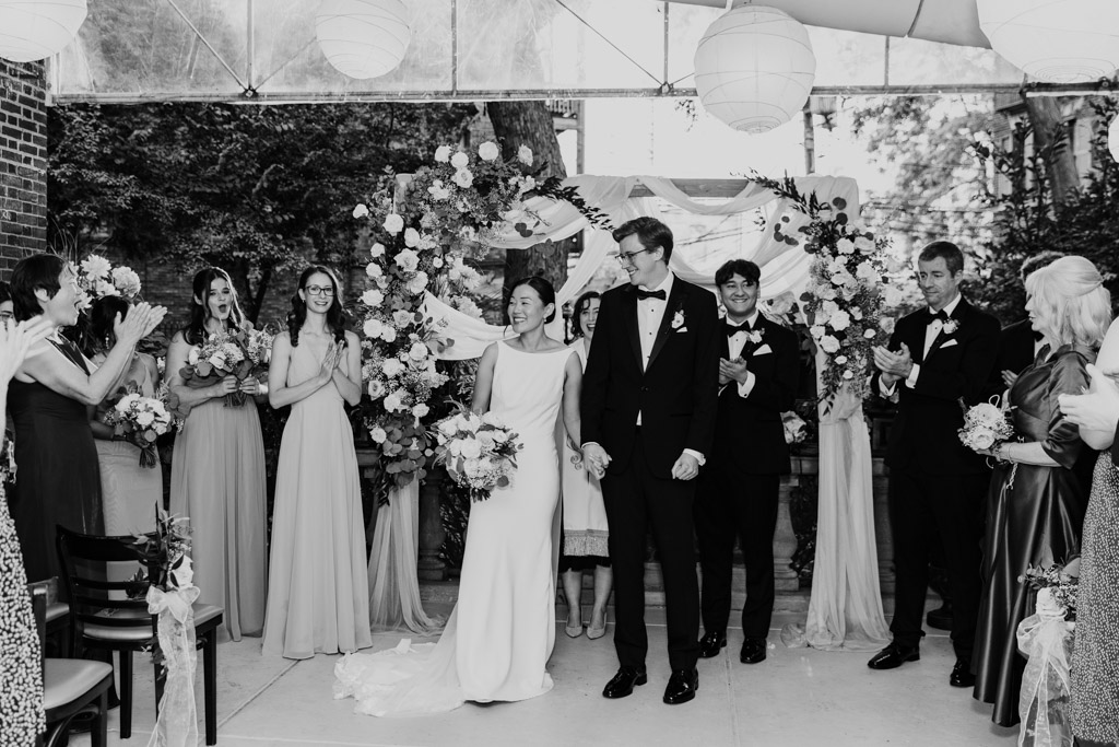 Black and white photo of newlywed couple holding hands and smiles at their Salvatore's wedding ceremony, surrounded by cheering guests and flowers