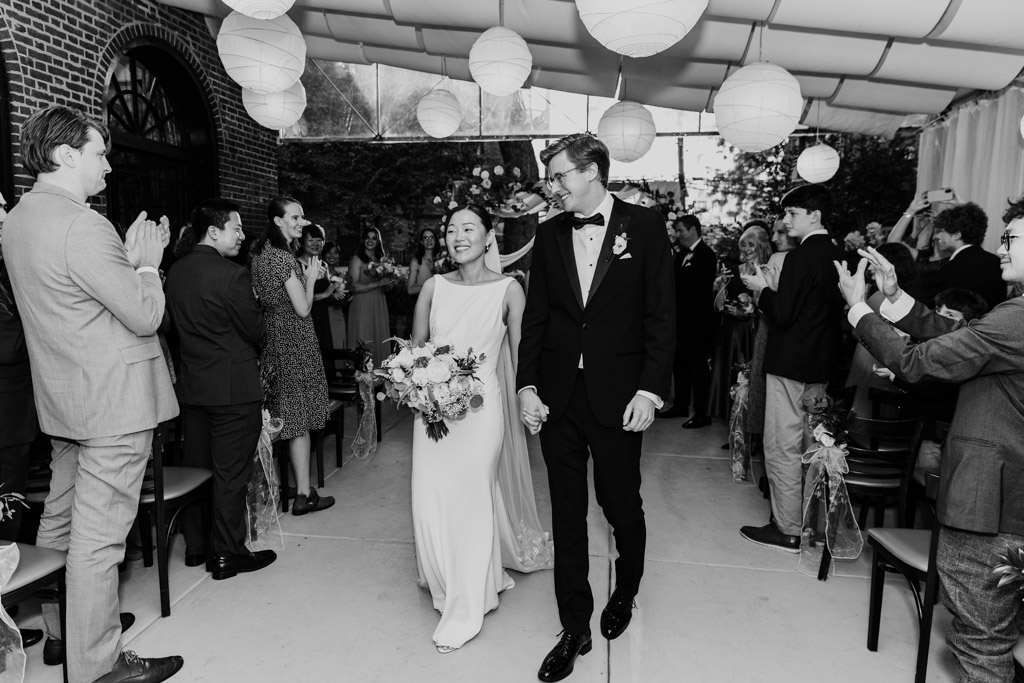 Black and white photo of newlywed couple walking down the aisle, smiling, as guests applaud at their elegant indoor wedding ceremony at Salvatore's