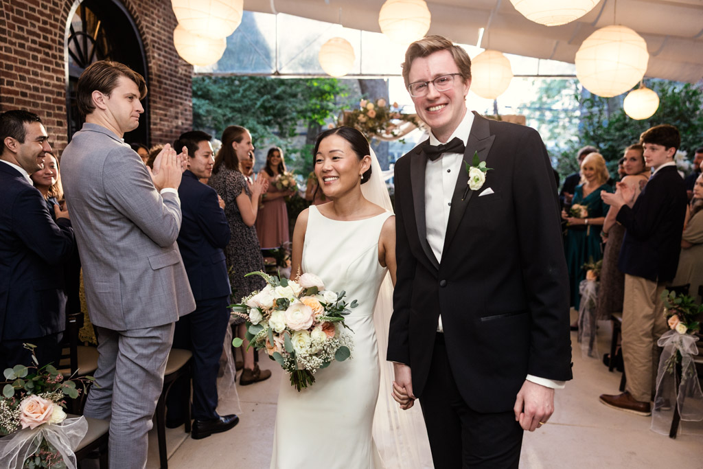 Bride and groom smile and hold hands while walking down the aisle at Salvatore's as guests applaud after their wedding ceremony