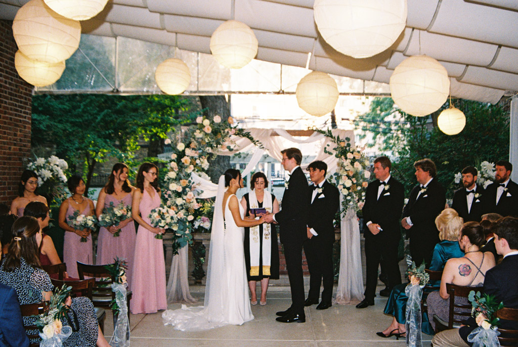 Film photo of bride and groom standing at the front of their wedding ceremony at Salvatore's, surrounded by their wedding party and guests, under glowing white lanterns