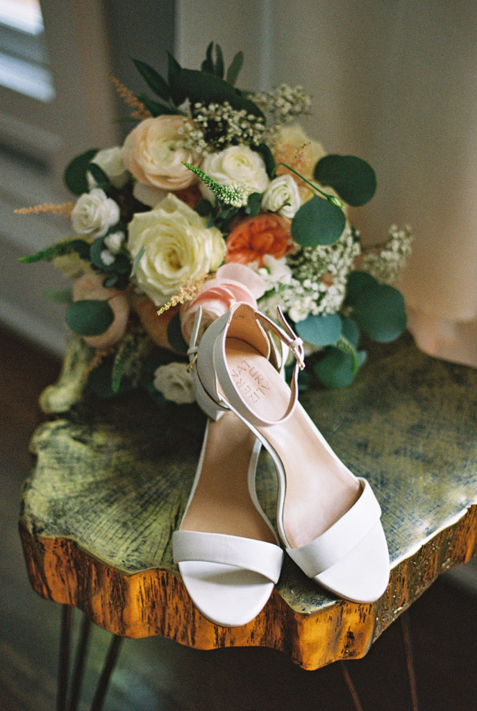 Film photo of bride's white shoes on a rustic table with a bouquet of pastel roses and greenery behind them