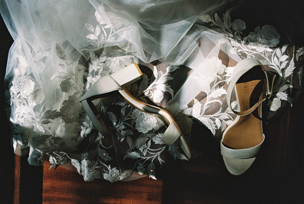 Film photo of bride's white shoes on a floral lace wedding dress, with sunlight highlighting the delicate textures