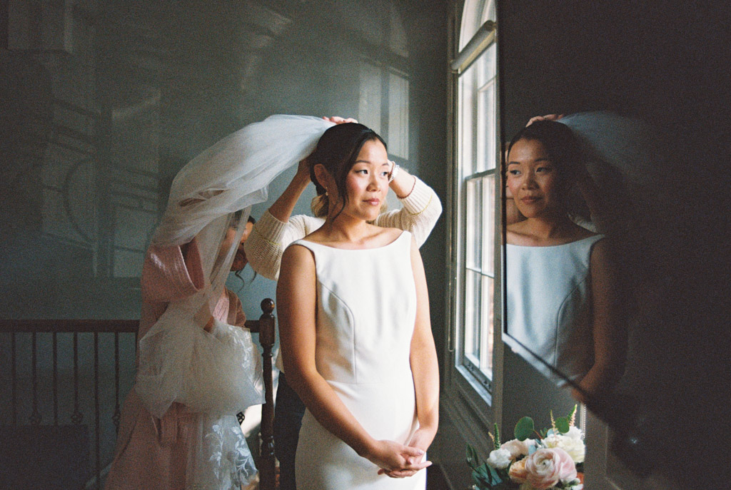 Creative film photo of bride standing by a window while someone adjusts her veil with her reflection visible in a nearby surface