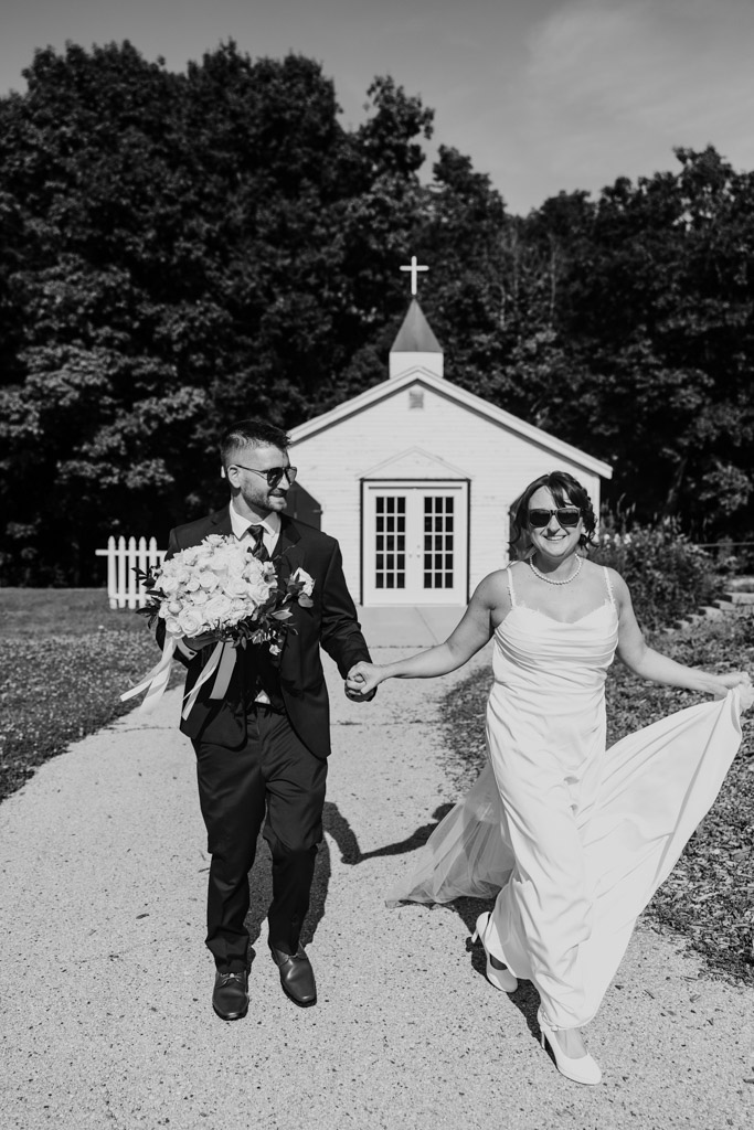 Black and white photo of bride and groom holding hands and smiling as they walk outside a small white chapel