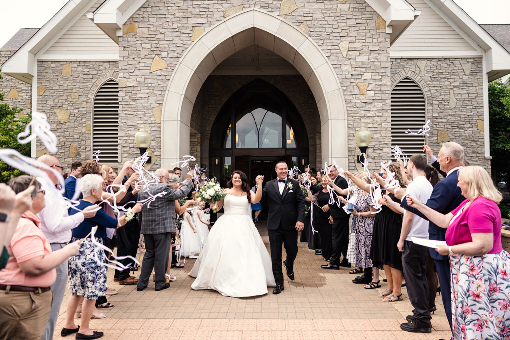 Bride and groom exit St. Katherine Drexel Church as guests celebrate and wave streamers on either side of them