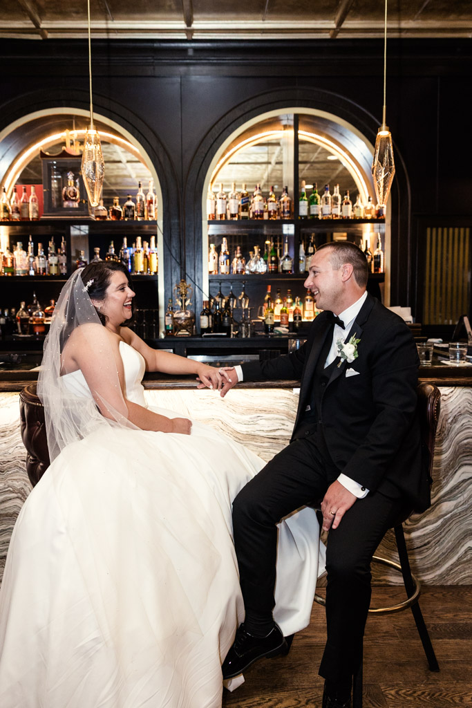 Bride and groom sit at a bar, holding hands and smiling at each other on their wedding day before their wedding reception at The Drake Oakbrook