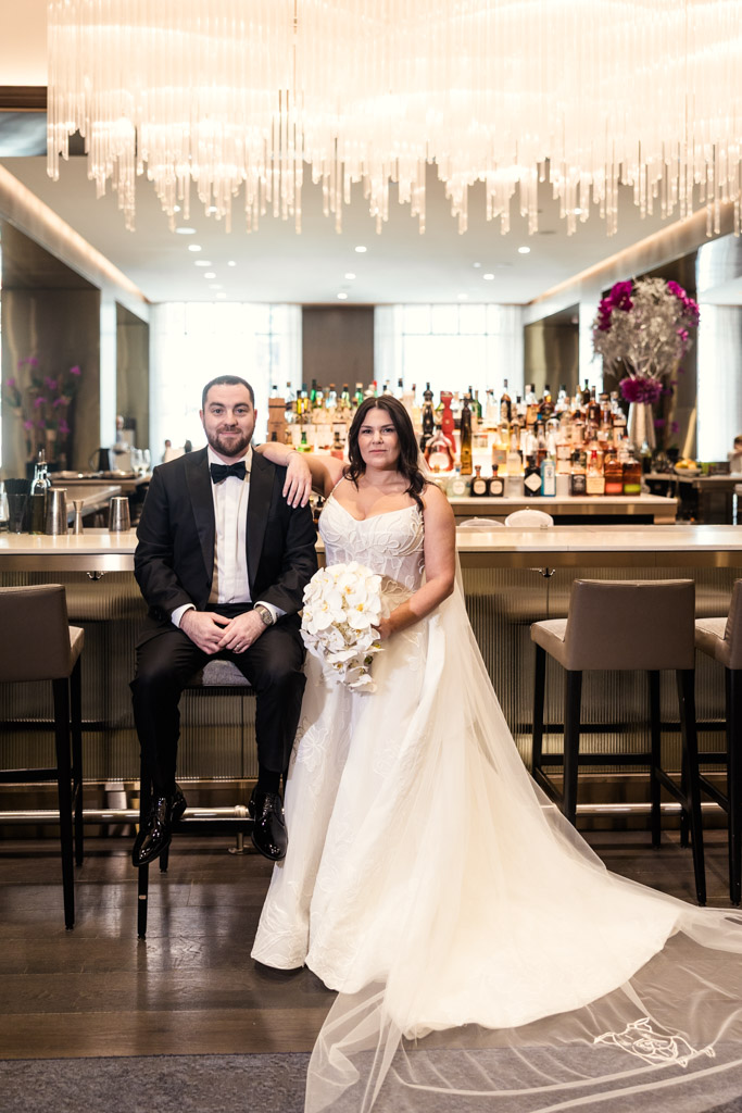 Portrait of bride and groom pose together at the stylish bar of The Four Seasons Chicago, the bride holding a white bouquet beneath a sparkling chandelier